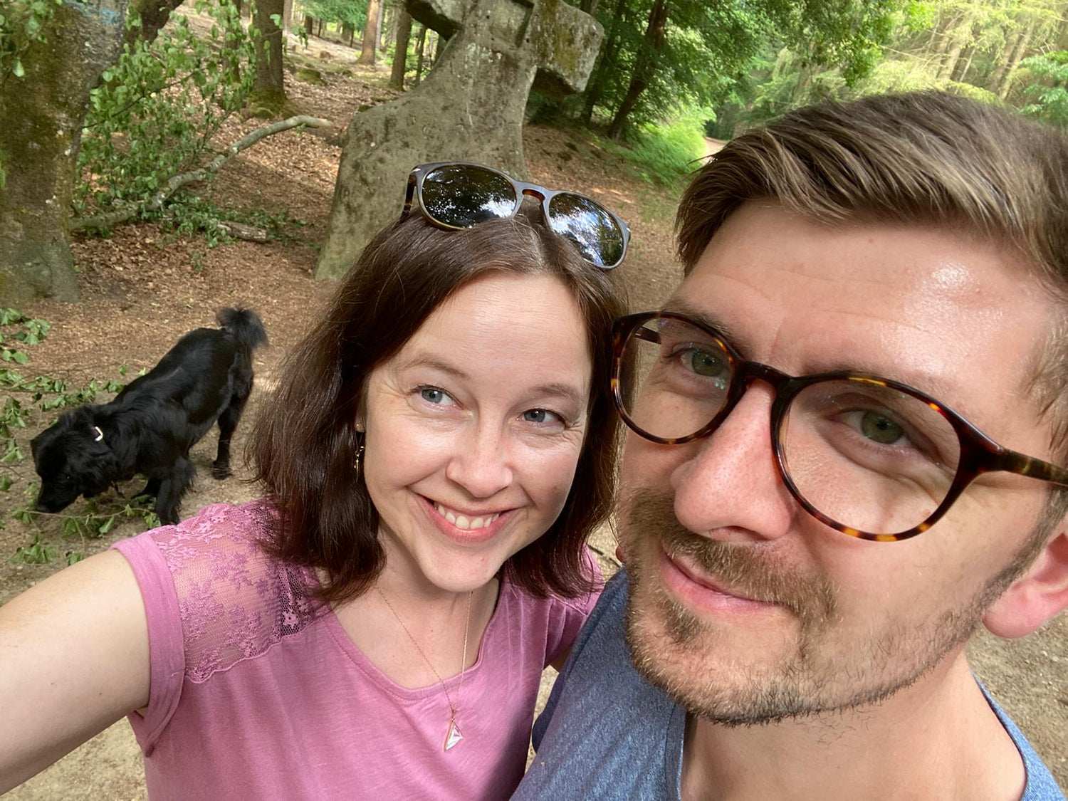 Two people taking a selfie in a forest with a black dog in the background.